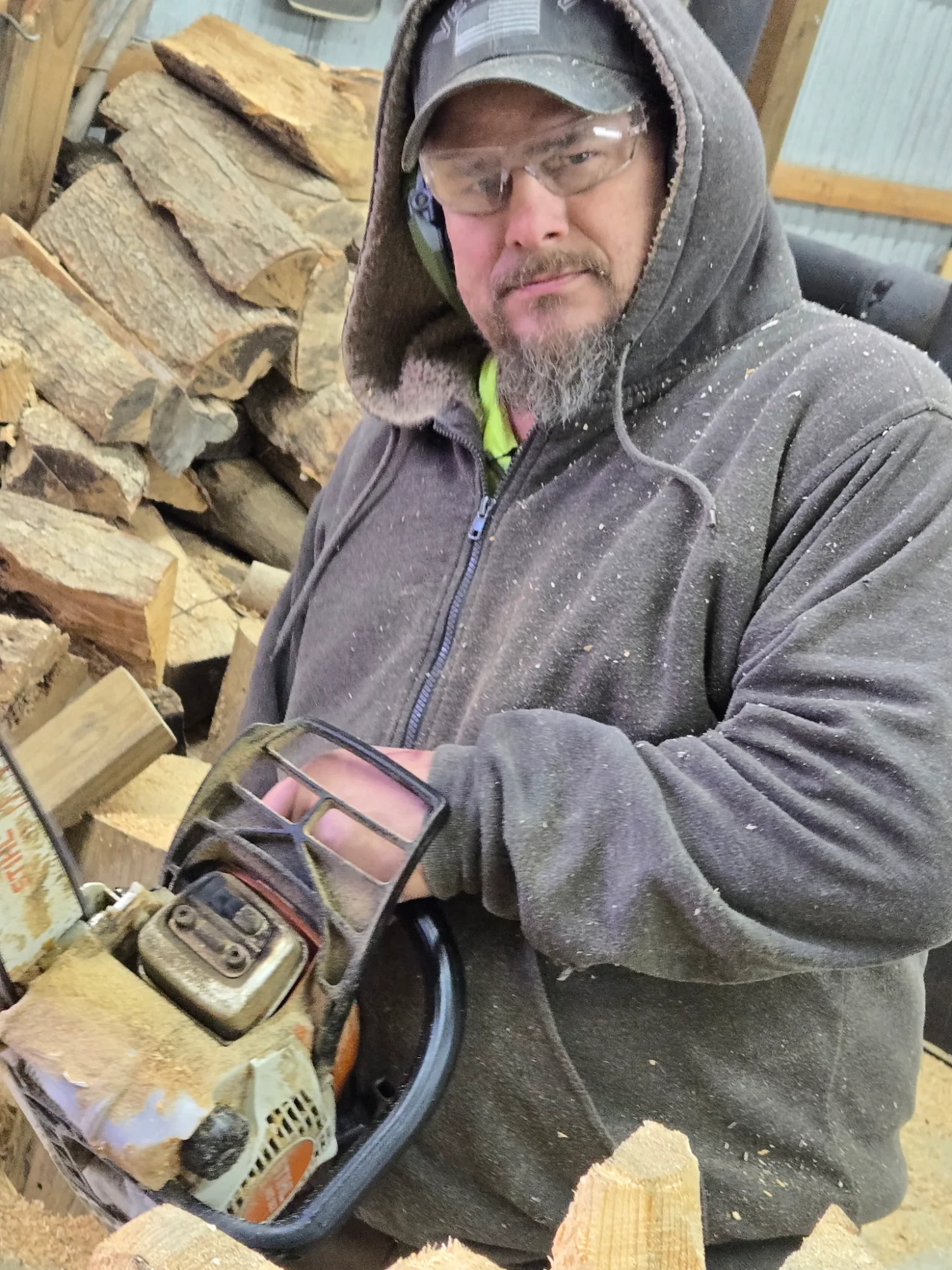 Gene holding a chainsaw with wood chips on his hoodie, ready to carve